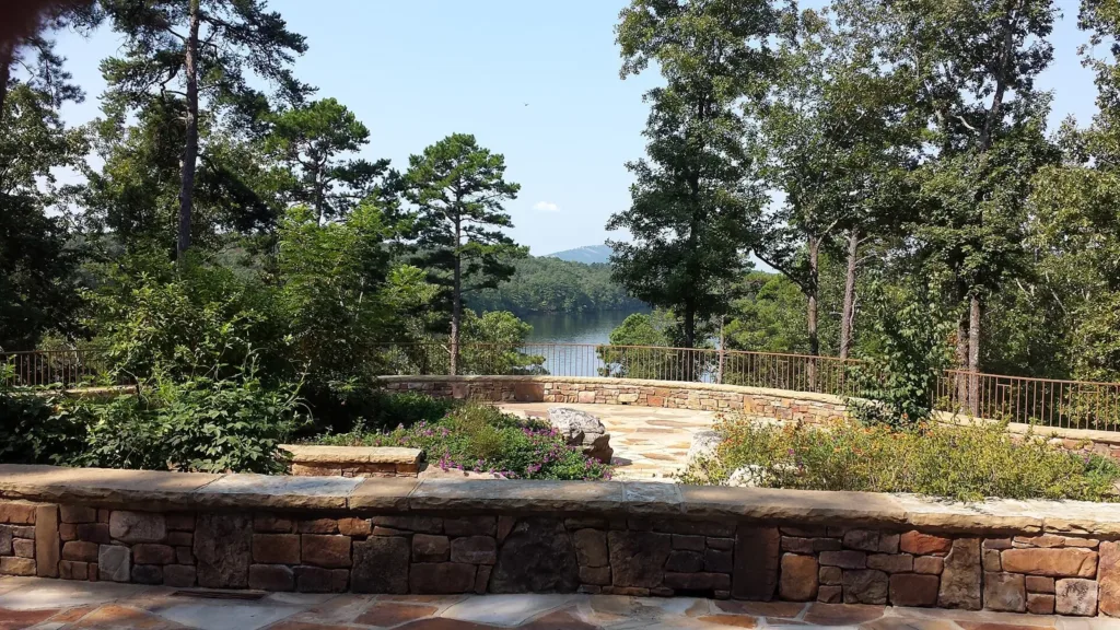 A curved stone terrace wall under tall pines, with water glinting through the trees beyond, at Garvan Woodland Gardens