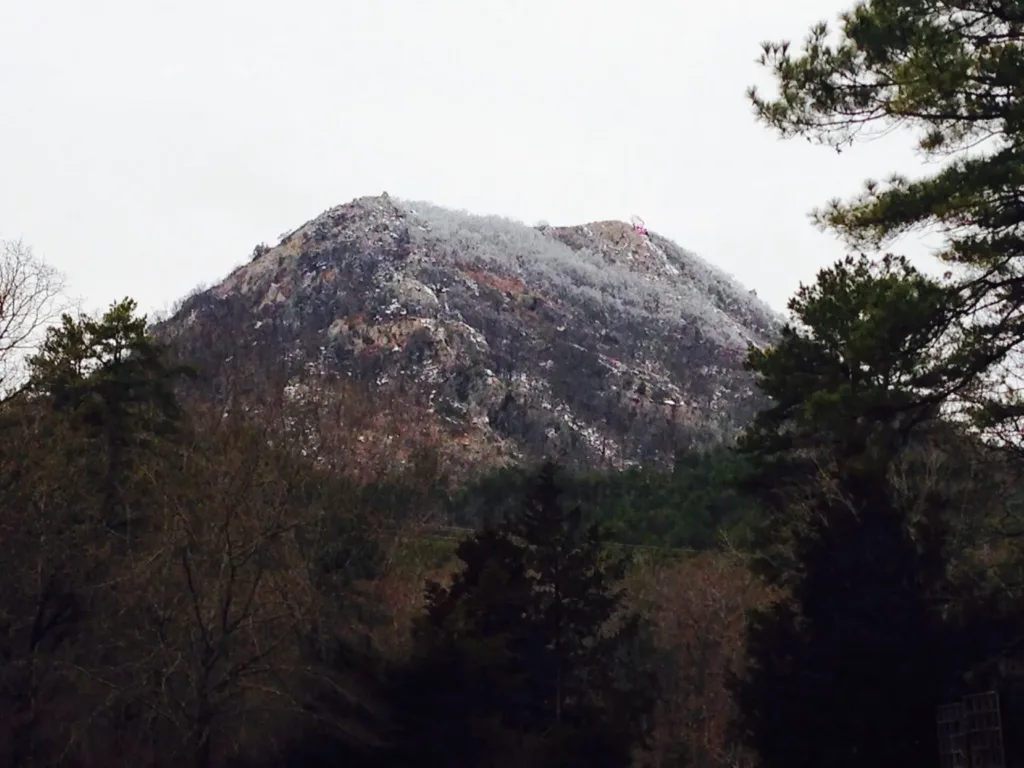 A rocky cone-shaped peak dusted with frost above a dark forest of pine trees
