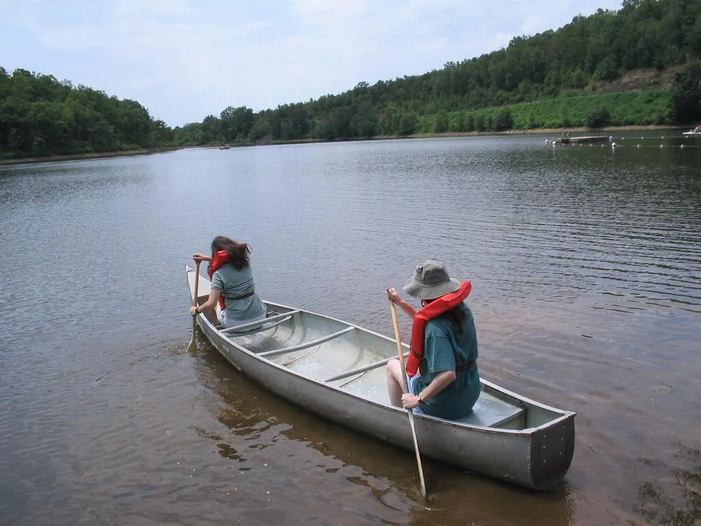 Two people in red life jackets paddling a silver canoe away from the shore across a calm lake ringed by green wooded hills
